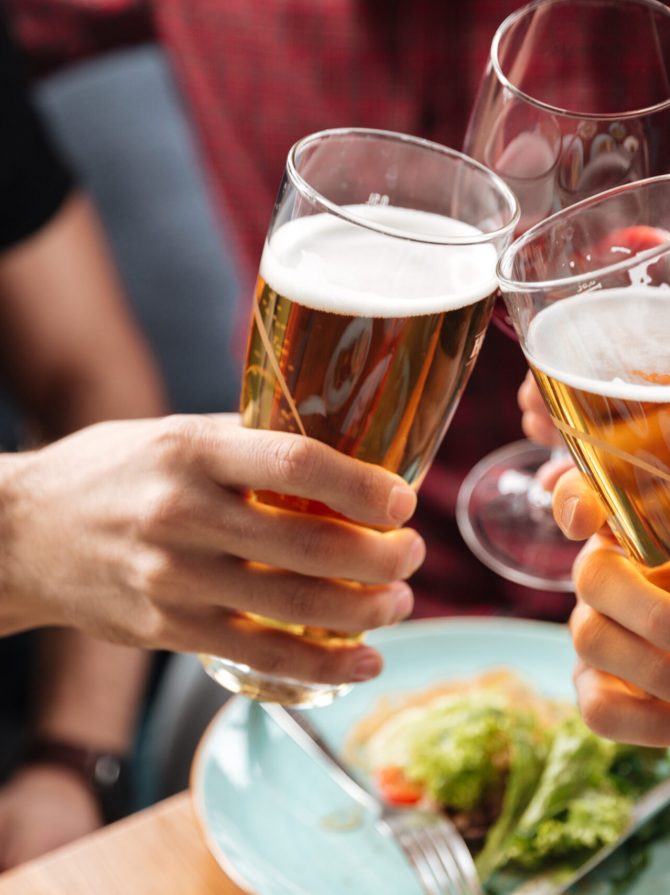 Cropped photo of young friends sitting in cafe while drinking alcohol. Focus on glasses of beer.